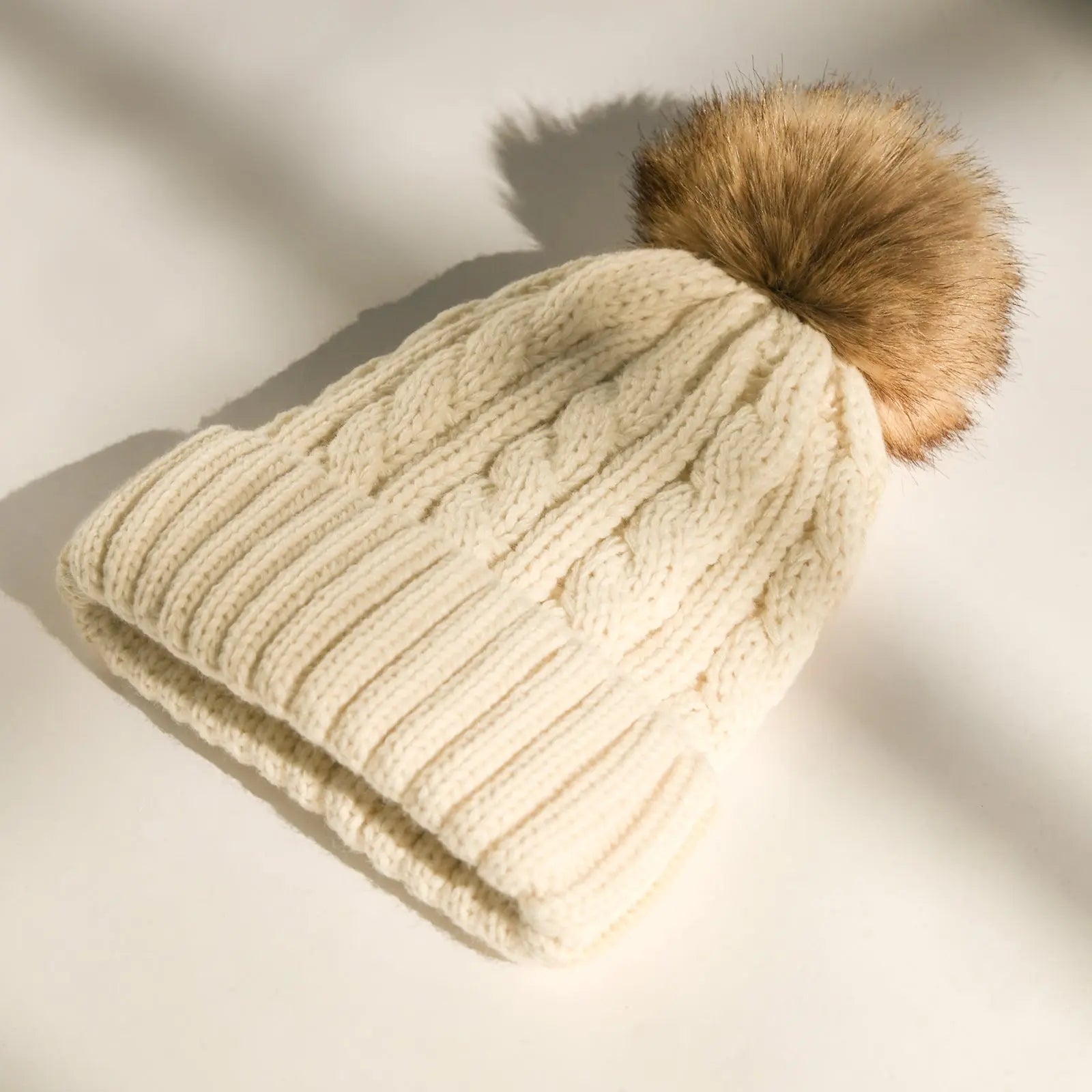 Cream cable knit winter beanie with brown faux fur pom, satin lined, on white background